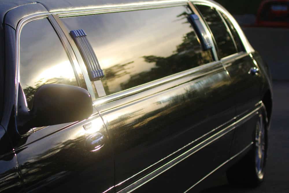 Close-up of a black limousine with tinted windows reflecting a sunset and trees, parked on a road.